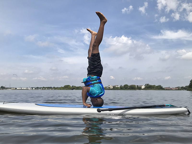 A Chill youth doing a headstand on a stand up paddle board.