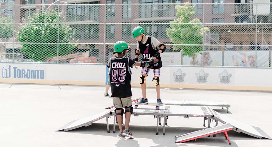 Chill youth holding another youths hand while he tries to drop in off a small skate ramp