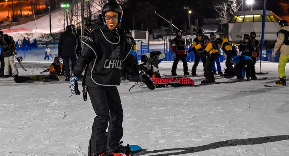 Chill youth standing on a snowboard facing down the mountain smiling at the camera thats looking uphill