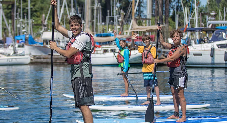 Chill youth paddling on SUP boards in a bay with boats in the background