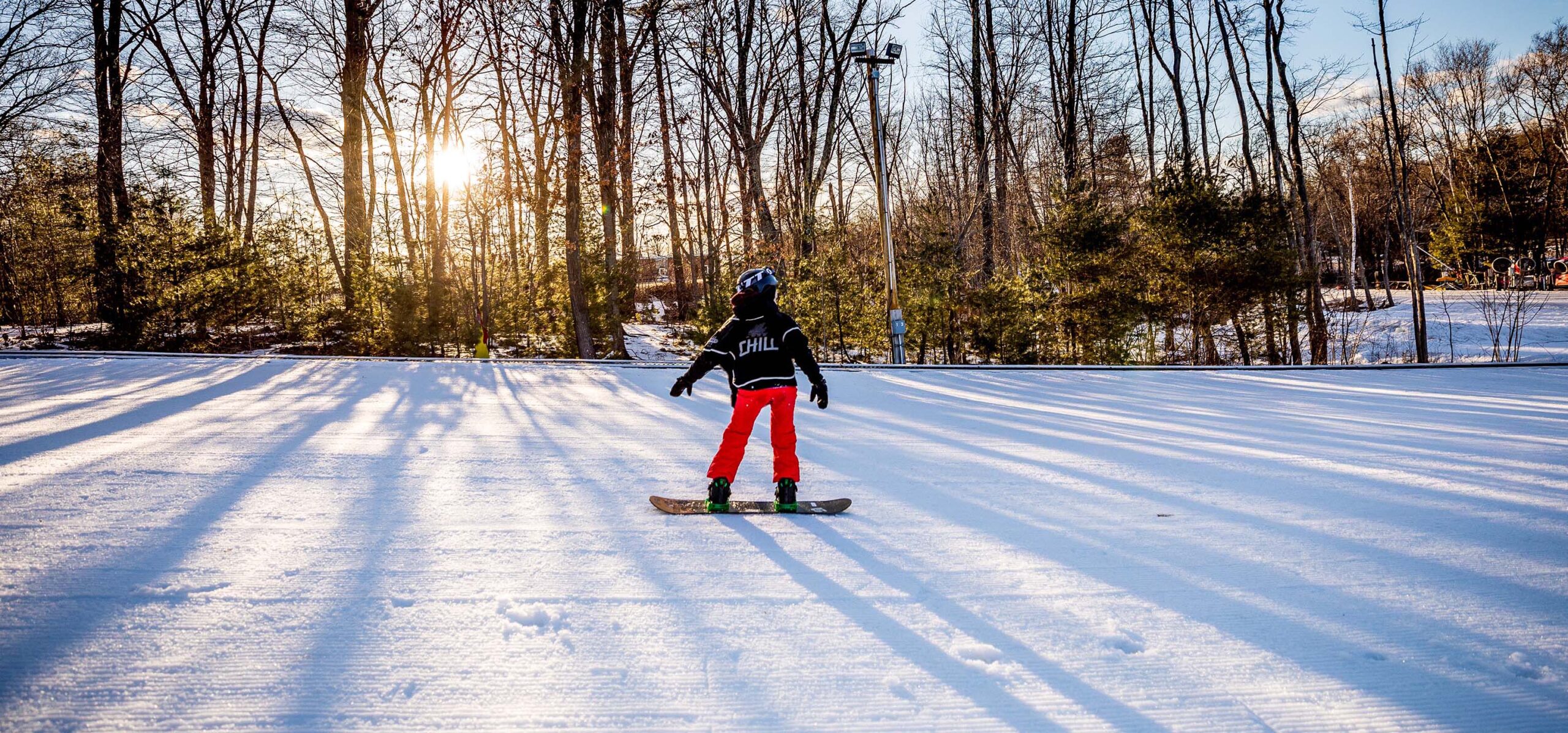A chill youth snowboarding with the setting sun shining through trees in the background.