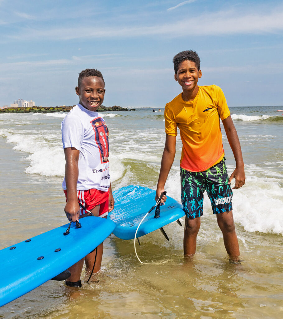 Two Chill youth smiling and holding surfboards in the edge of the water.